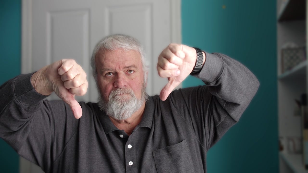 An older man communicates his feelings by using his hands to show a thumbs down. He sits in a well-lit room with blue walls. His expression shows his discontent.