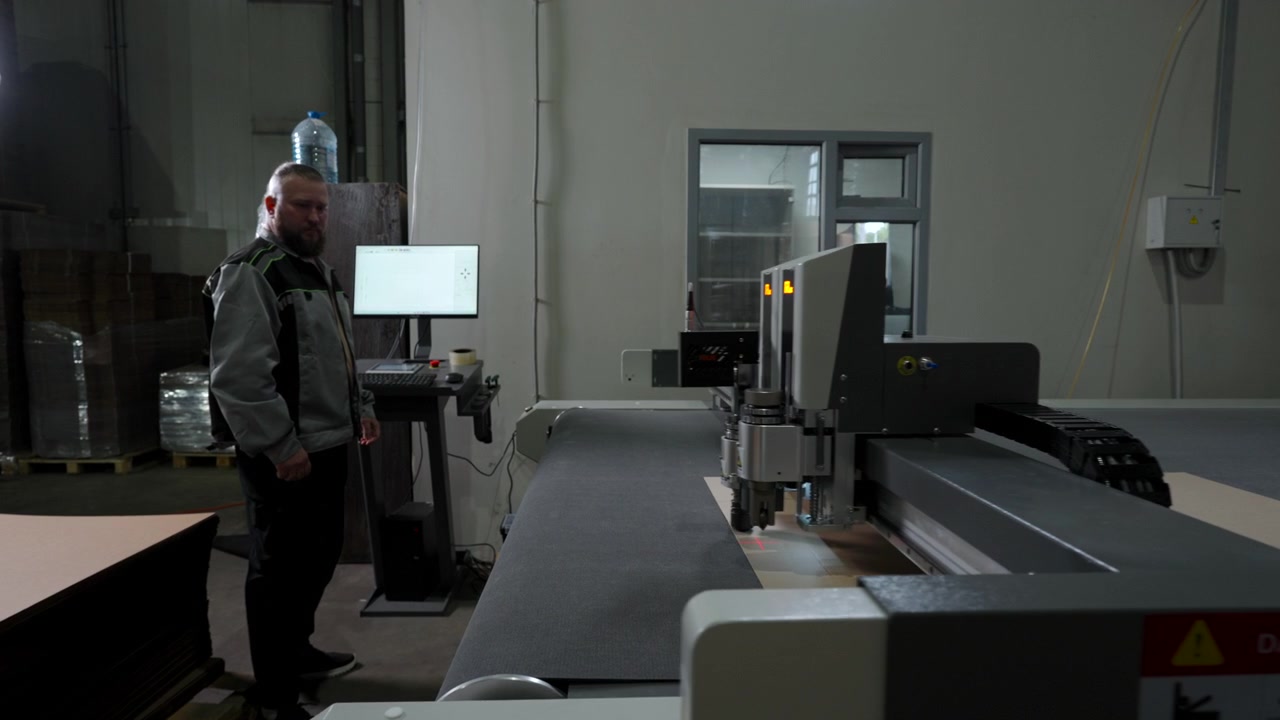 Adult man stands beside automated cutting machine inside dim industrial workshop, watching control screen near stacked materials, cables, pallets, and windowed wall