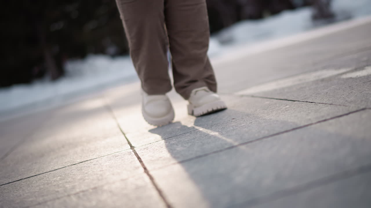Closeup of woman legs in beige corduroy pants and chunky white platform shoes tapping rhythmically on large tiled snowy plaza ground, casting long evening shadows under soft pale winter sky light