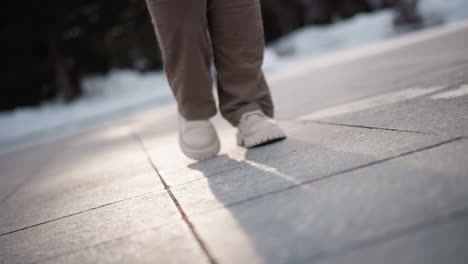 closeup of woman legs in beige corduroy pants and chunky white platform shoes tapping rhythmically on large tiled snowy plaza ground, casting long evening shadows under soft pale winter sky light
