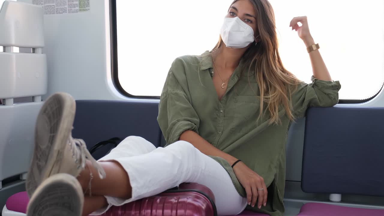 mujer joven con máscara facial con las piernas en su maleta viajando en tren. nuevo concepto de viaje normal.