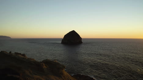 Aerial-ascend-above-sandstone-cliffs-to-haystack-rock-centered-at-sunset,-Oregon-Coast