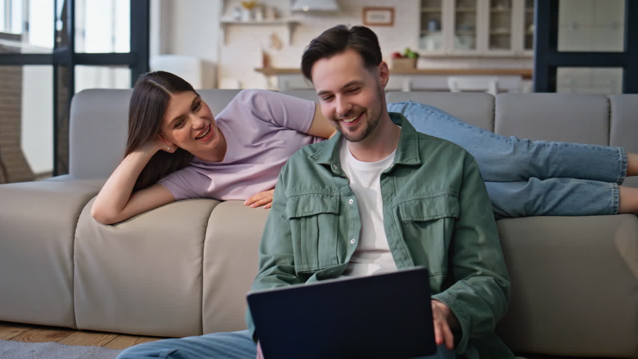 Smiling man working laptop lying girlfriend resting at sofa talking to husband