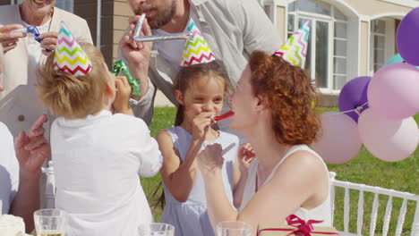 playful little brother and sister blowing party horns and having fun with parents and grandparents on outdoor celebration dinner on summer day