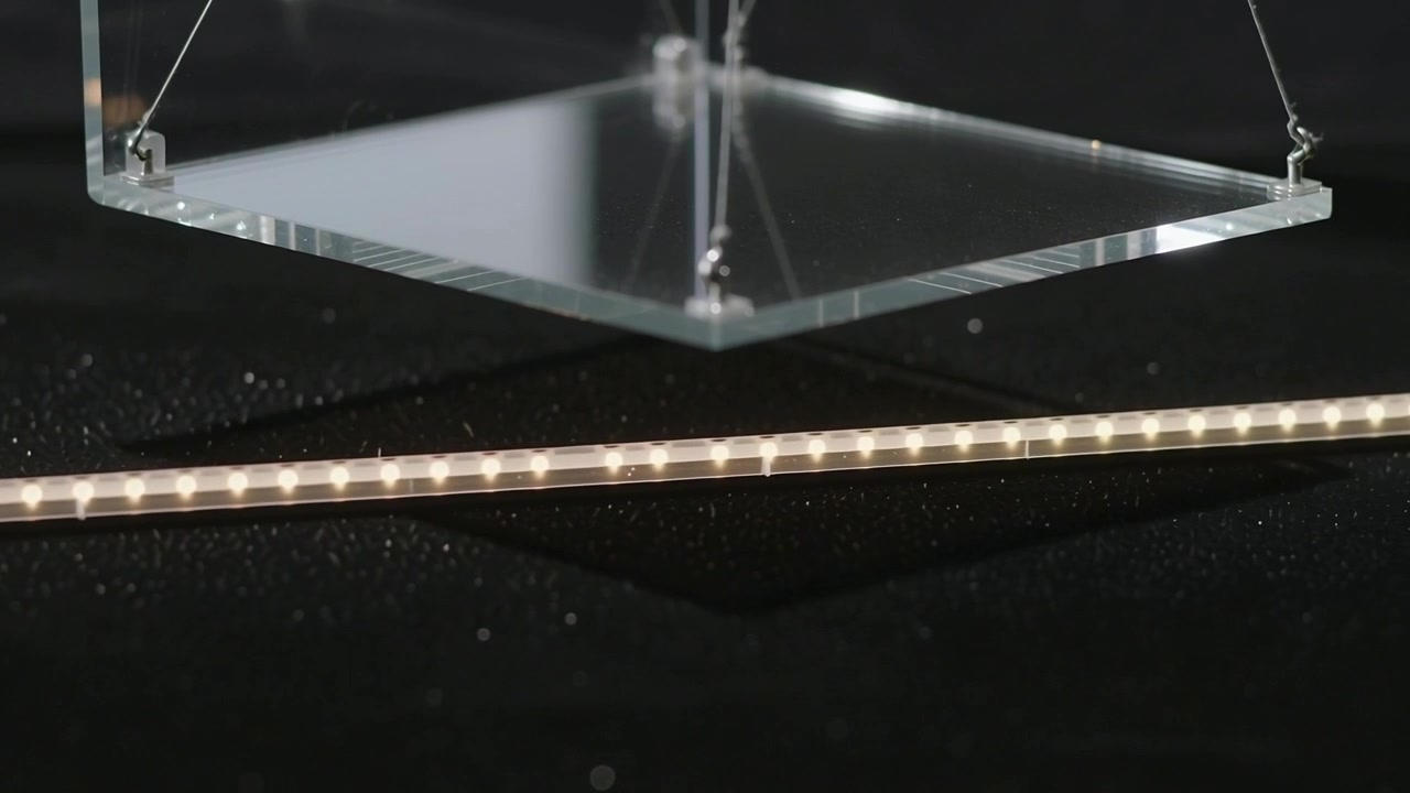 Equipo de cámara fuera de plano empujando un tubo LED delgado sobre una mesa de estudio, con un panel acrílico inclinándose