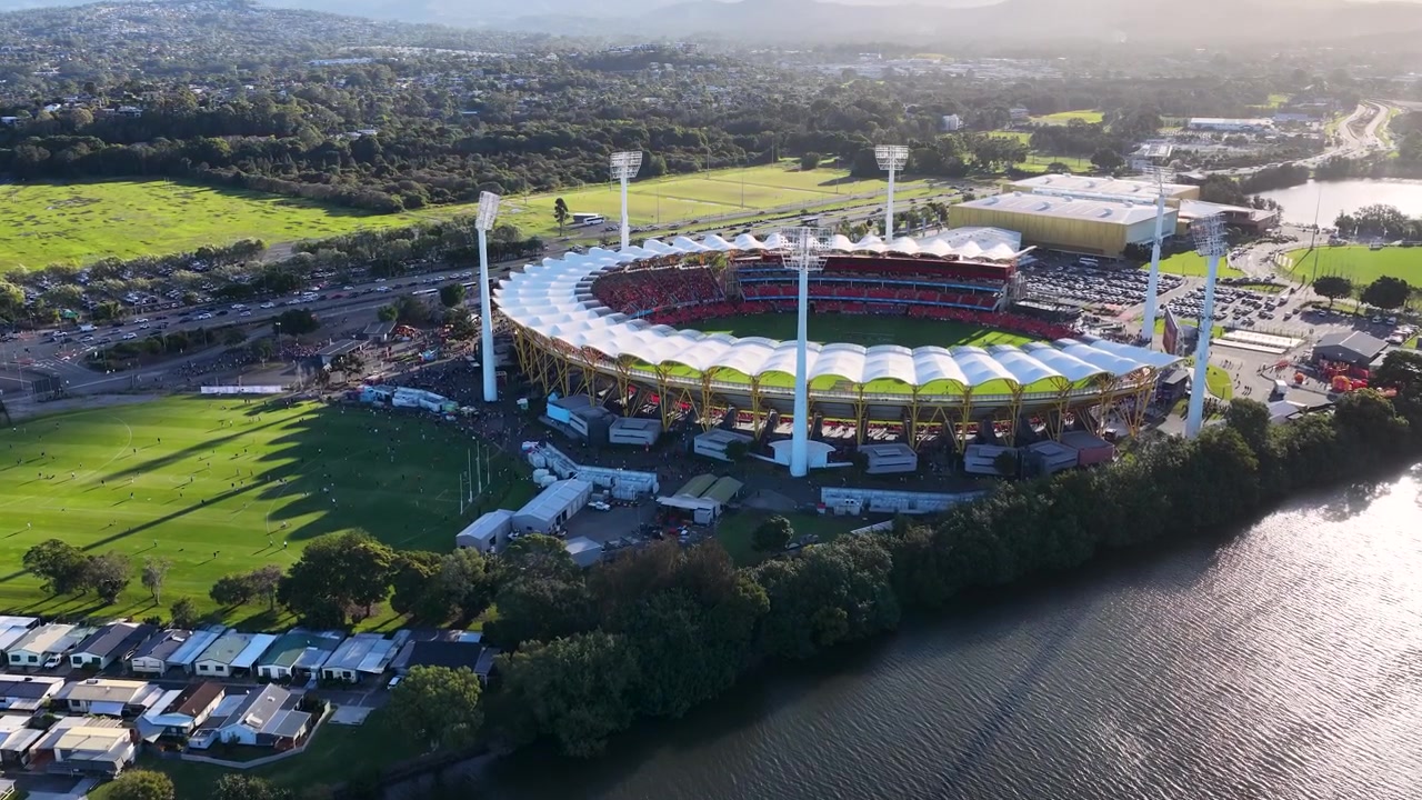 Drone footage captures a large circular sports arena near a river during golden hour.