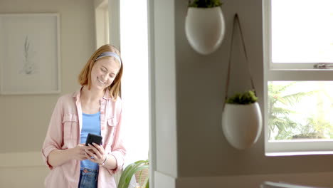 smiling woman using smartphone while standing near window at home