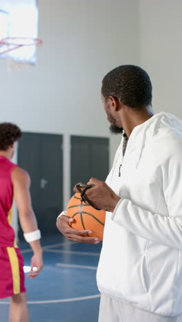 vertical video: holding basketball, coach observing player practicing in school gymnasium