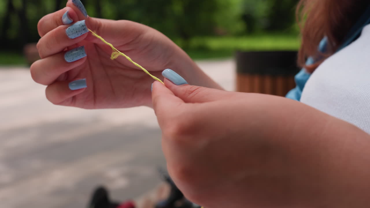 Close up hands separating green embroidery thread outdoors, blue nails gently pulling strand against blurred park background, careful craft preparation before stitching