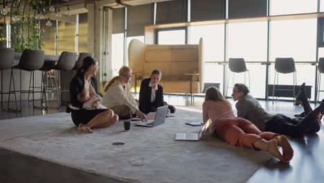 Vista-Lateral-De-Un-Grupo-De-Mujeres-De-Negocios-Vestidas-Con-Ropa-De-Negocios-Acostadas-En-La-Alfombra-En-Una-Oficina-Moderna-Hablando-Y-Durante-Un-Descanso-Entre-El-Trabajo,-Una-Niña-Morena-Amamanta-A-Su-Pequeño-Hijo-En-La-Oficina.