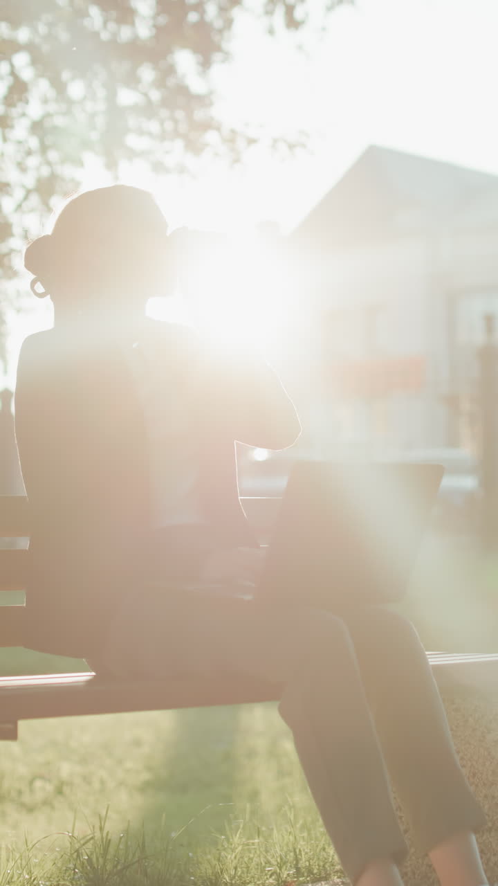 Silhouette effect surrounds woman working outdoors in park while sipping beverage from cup with laptop on lap and blurred background featuring buildings, trees, and cars
