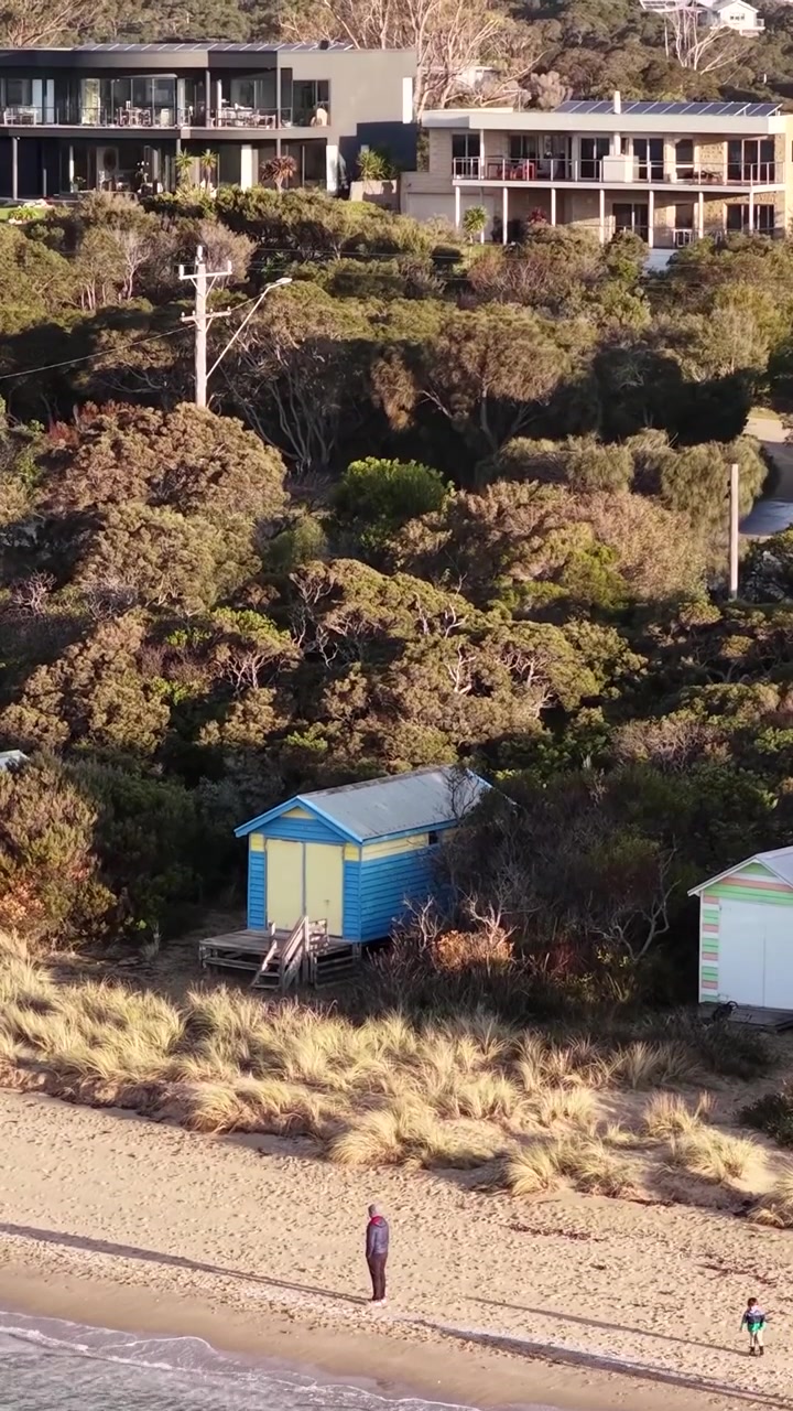 Et vertikalt dronebillede fanger en blå badekabine indlejret i kystvegetation ved solnedgang.