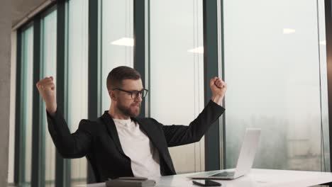 A-happy-man-in-business-clothes-in-the-office-looks-at-the-laptop-screen,-raising-his-hands