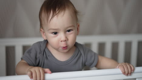 little adorable baby boy standing in his crib after waking up. cute toddler making funny faces in front of camera. close up.
