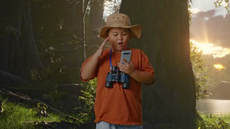 niño asiático con sombrero y binoculares mirando su teléfono inteligente y luego diciendo wow. niño investigador examina algo mientras explora la naturaleza del bosque, el concepto de aventura de turismo de viaje