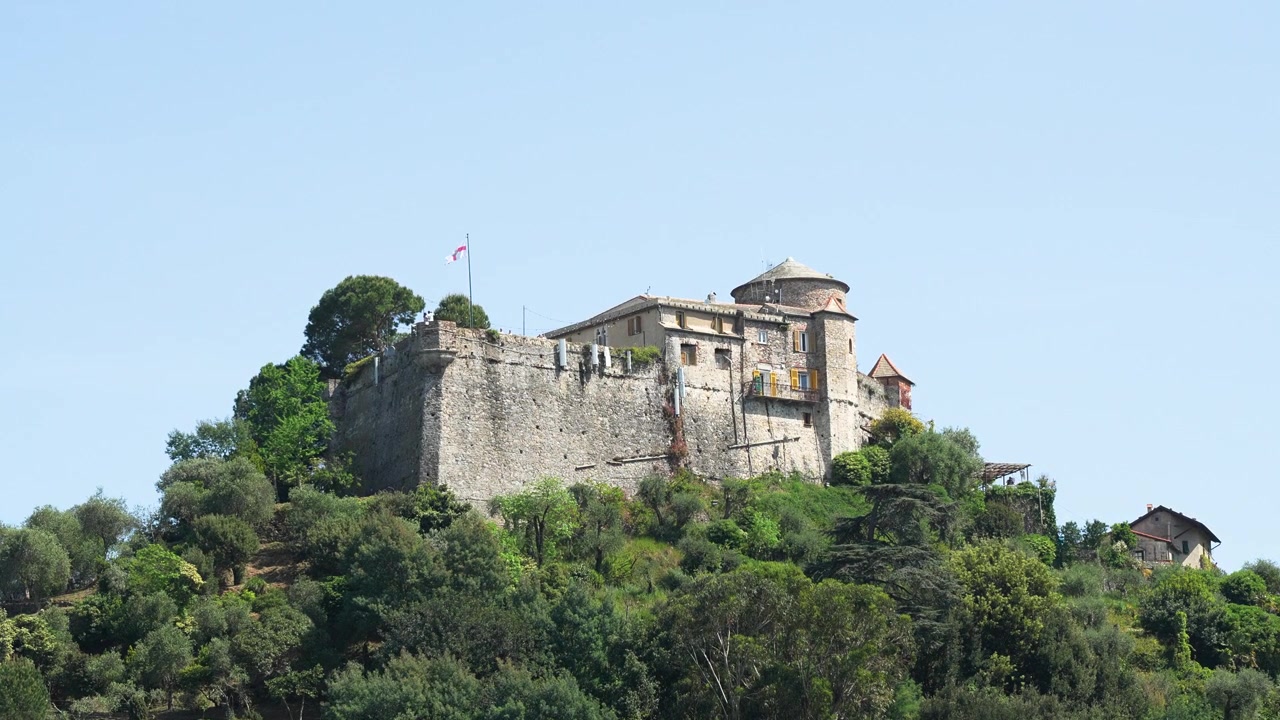 Riprese con drone mostrano Castello Brown arroccato su colline verdeggianti a Portofino, Italia, sotto cieli azzurri e sereni. La fortezza storica è incorniciata da alberi mediterranei e una luce diurna soffusa.