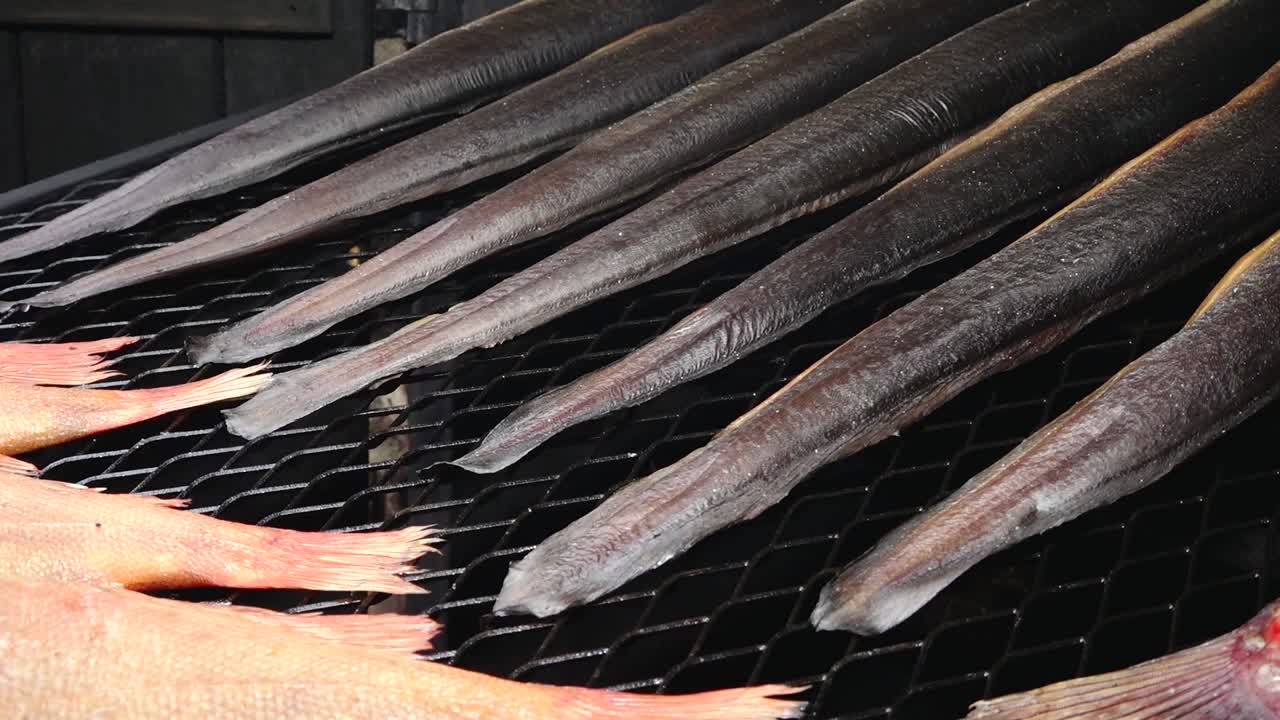 Various types of fish, including eel and redfish, are being smoked on a grill rack. The golden-brown color and rich texture highlight the traditional smoking process.