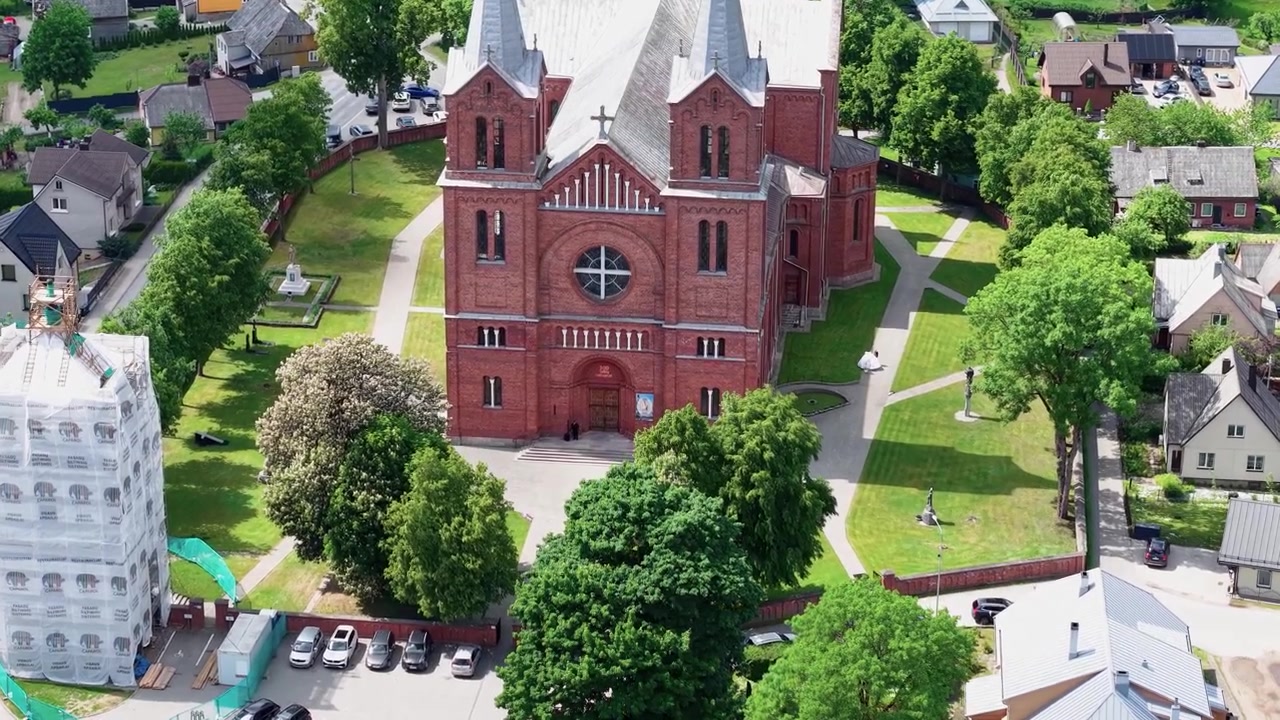 Imagem de drone mostra a Igreja de São João Batista de tijolos vermelhos em Plungė, cercada por árvores verdes exuberantes, gramados bem cuidados e edifícios históricos da cidade sob a luz natural brilhante do dia