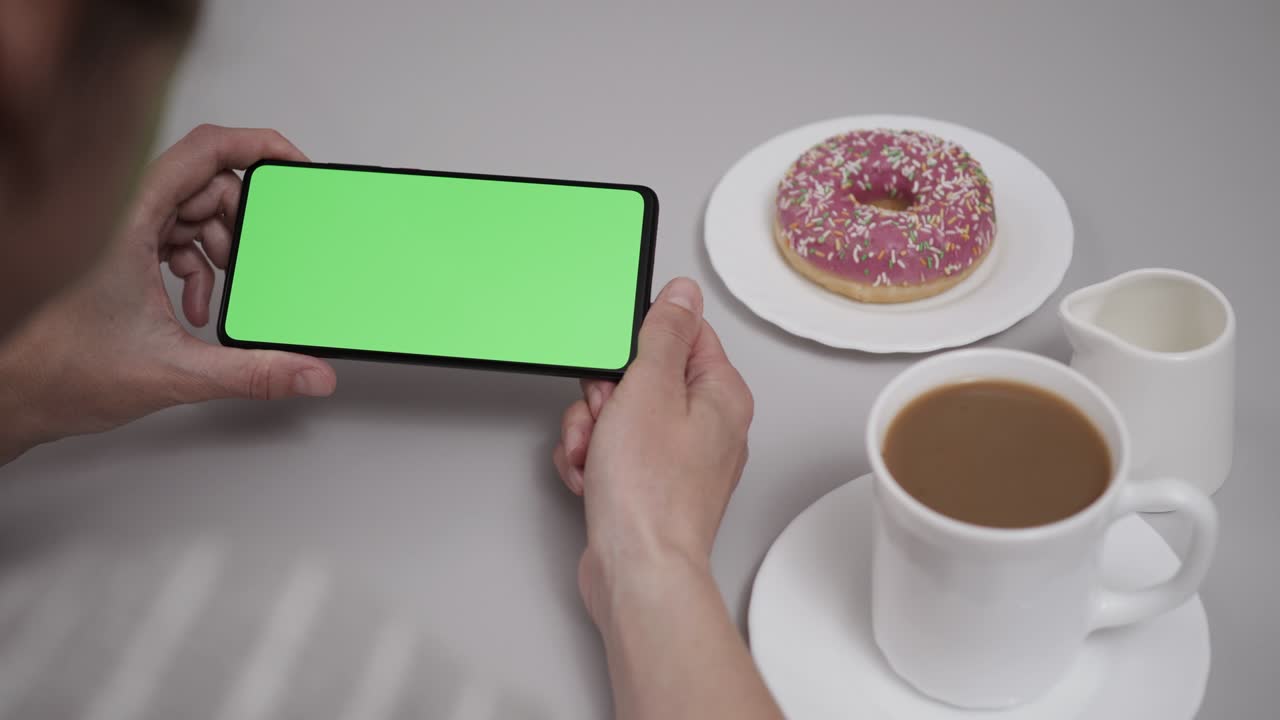 mujer sentada en la mesa de café donut usando un teléfono inteligente con pantalla verde de clave de croma, desplazándose a través de las redes sociales medios de comunicación en línea tienda internet. teléfono inteligente en modo horizontal con maqueta de pantalla verde.