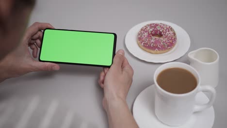 woman sitting at table coffee donut using smartphone with chroma key green screen, scrolling through social network media online shop internet. smartphone in horizontal mode with green screen mock-up.