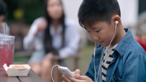 asian child listening music and typing on smartphone in outdoor cafe