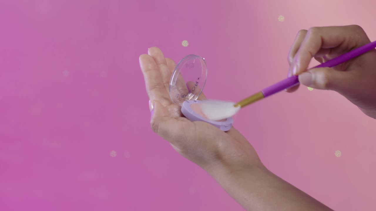 Close up of a young woman's hand picking up blush with a fan brush, make up, cosmetics, on a moving pink background