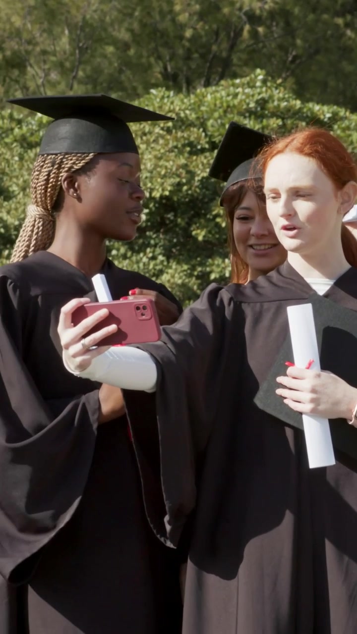 Video verticale: Un laureato si sistema il cappello, guida altri laureati in toga che posano su una terrazza con i diplomi per una foto scattata con il telefono.