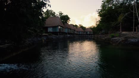 Aerial-view-over-a-river-flowing-through-luxury-beach-wooden-houses-in-a-tropical-resort-surrounded-by-trees,-in-Tulum,-Mexico,-at-sunset