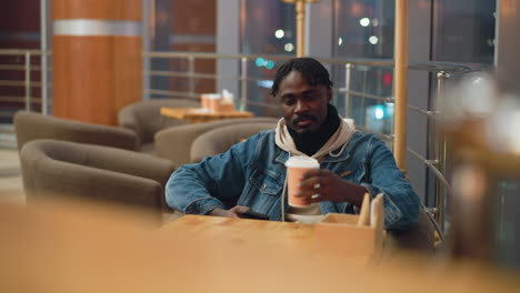 young man in denim jacket and beige hoodie seated alone in warm modern cafe at night, holding takeaway coffee cup and smartphone, looking calm and reflective with soft indoor lights in background