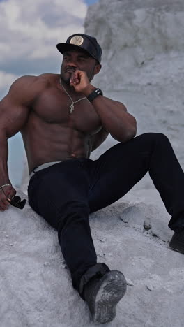 thoughtful black skinned guy sitting on white stone. shirtless african american man with muscular body rests outdoors. sportsman on hill in summer. vertical video