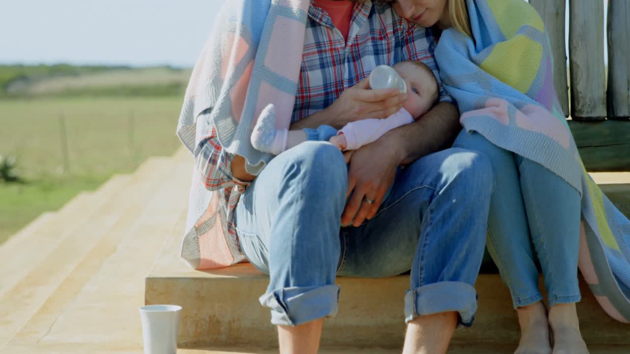 vista frontal de padres caucásicos adultos alimentando a su bebé con una botella en el patio trasero de la casa 4k
