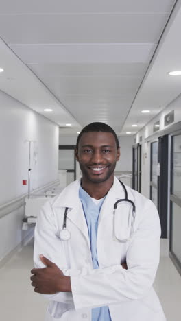 vertical video of portrait of happy african american male doctor in hospital corridor, slow motion