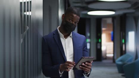 Portrait-of-african-american-businessman-wearing-face-mask-using-tablet-in-modern-office