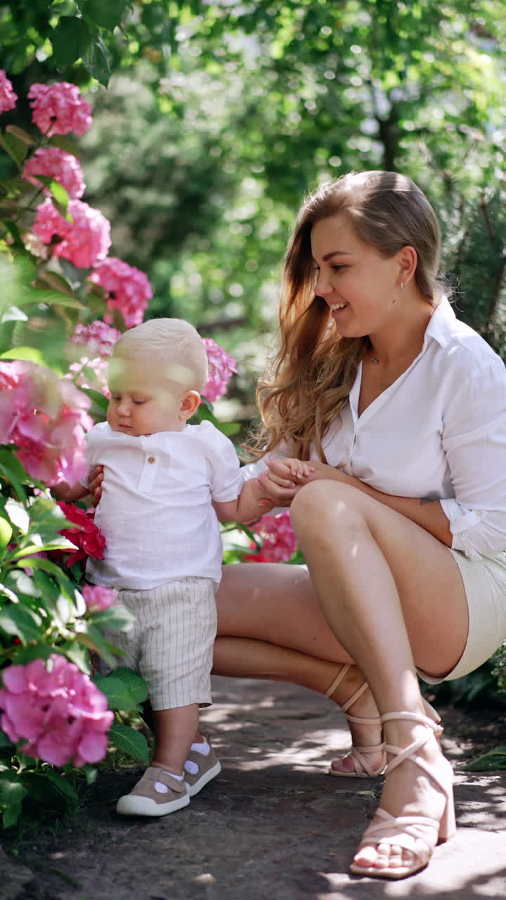 Happy mother and her little baby son near the blossoming bush. Cute kid touches the flowers happily. Vertical video.
