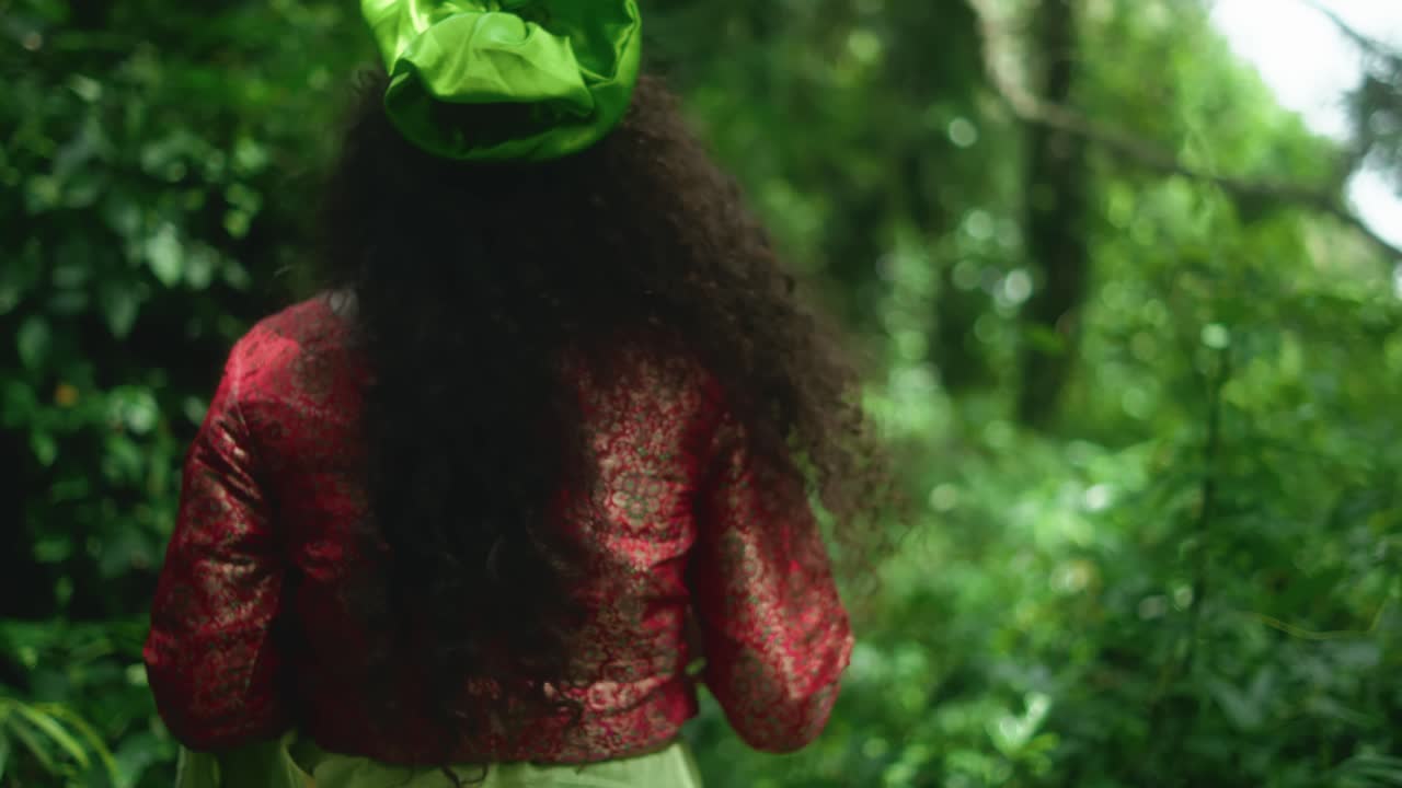 A woman faces the camera holding a leafy branch, then turns and walks through the jungle. She wears a red dupatta, a green fabric, and has a flower in her hair