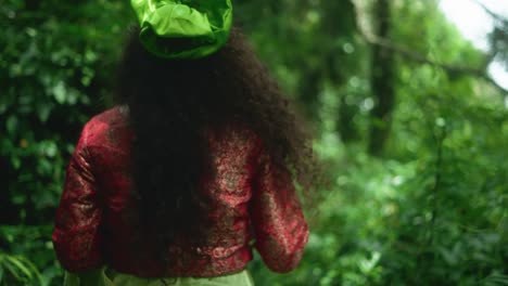 a woman faces the camera holding a leafy branch, then turns and walks through the jungle. she wears a red dupatta, a green fabric, and has a flower in her hair