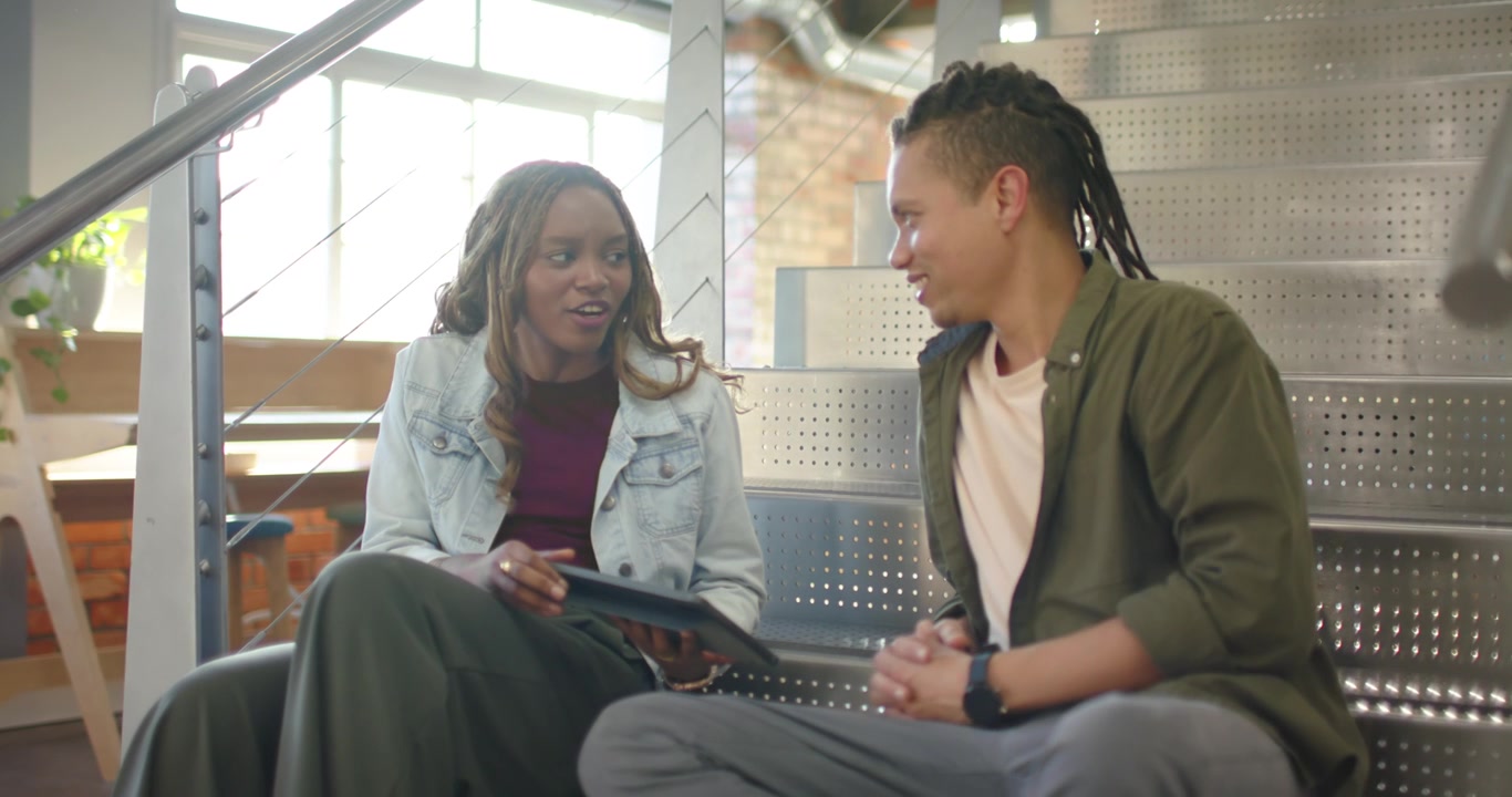 African American coworkers sitting on stairs, woman tapping tablet, man reviewing tablet content