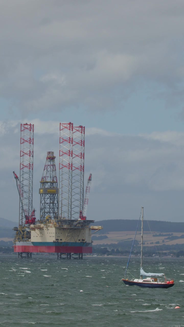 En lille sejlbåd bevæger sig over oprørt hav foran en stationær offshore olieplatform under overskyet dagslys, med fjerne bakker i Cromarty, Skotland