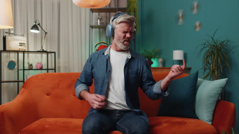 Adult-man-in-headphones-dancing-on-sofa-at-home-listening-favorite-music-playing-on-imaginary-guitar