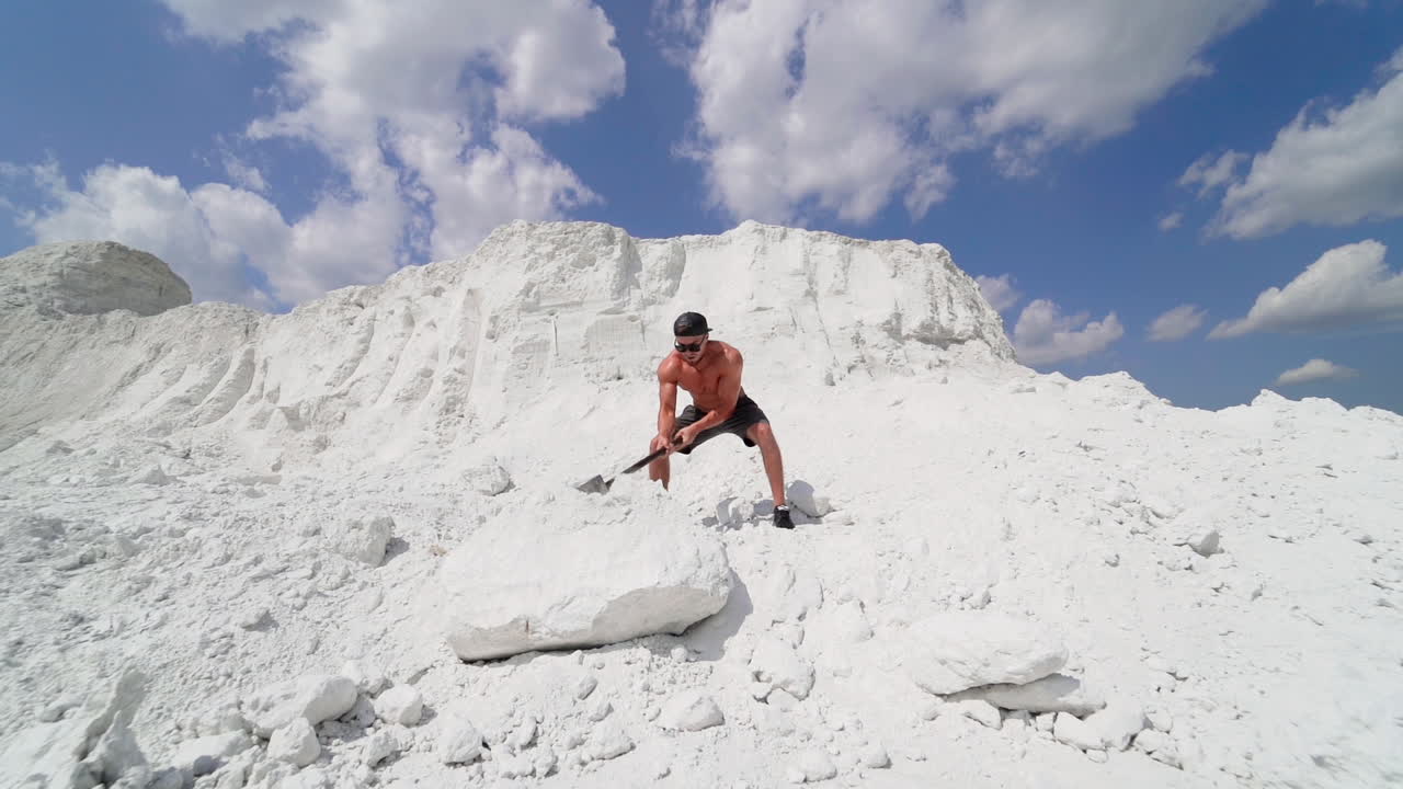 Sportsman with huge hammer outdoors. Sporty man demolishing big white rock by metal tool on the top of mountain in summer day. Slow motion.