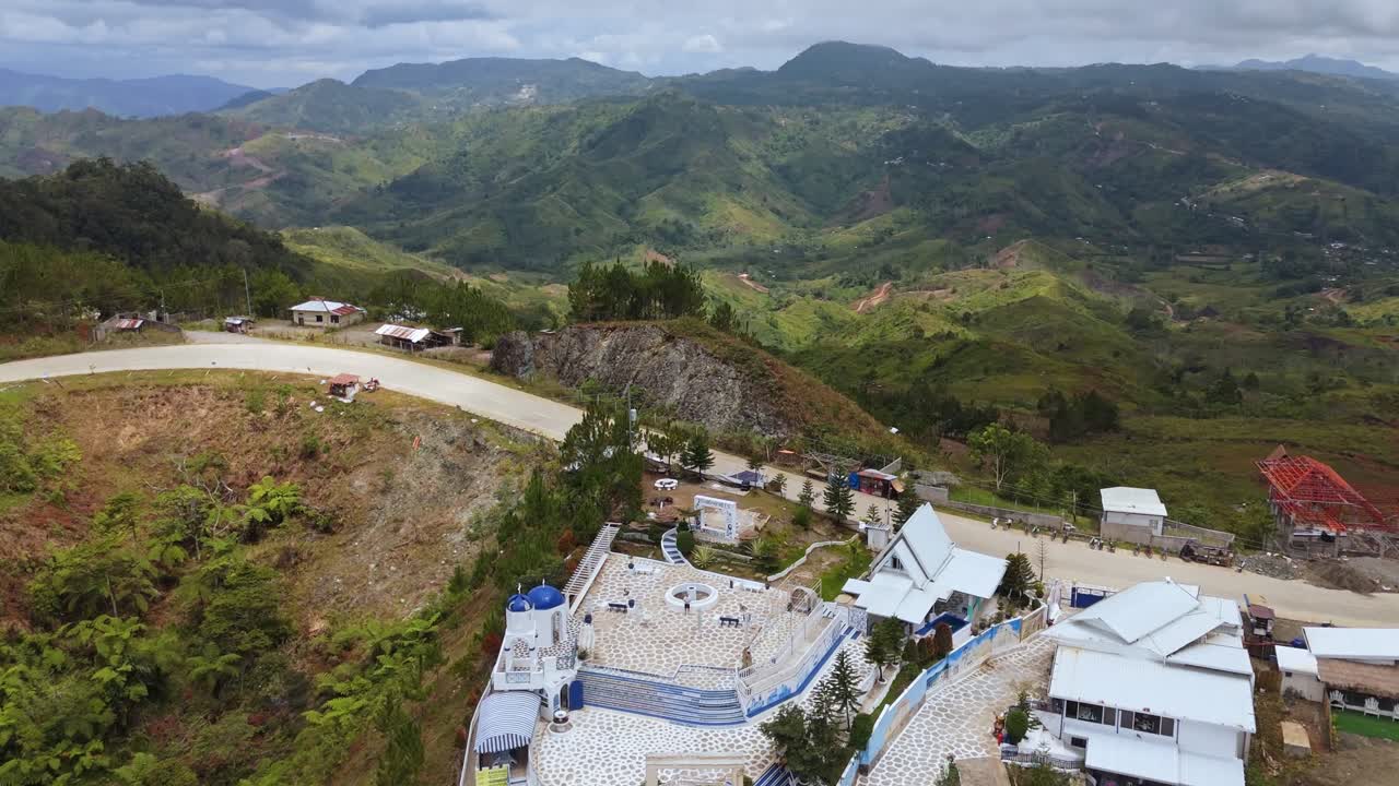 Aerial view of a winding mountain road and hillside village surrounded by lush green valleys in the Philippines. Scenic rural landscape with homes, hills, and cloudy skies captured in daylight