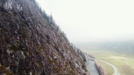 Moody-foggy-steep-mountain-slope,-fields-with-farm-houses-and-a-street-in-distance