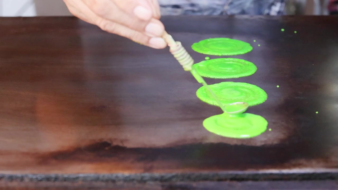 Close-up shot of a vendor spreading green batter into thin pancakes on a flat griddle.