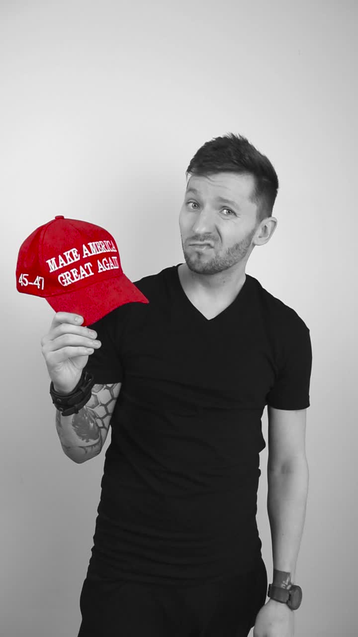 Grayscale footage of a man with tattoos taking off a red colored MAGA Make America Great Again hat, looking at it and begins shaking his head with disapproval and disgust against Trump, unhappy man.