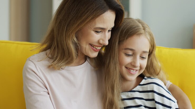 Close Up View Of Blonde Mother And Daughter Smiling And Watching Something On Tablet While Sitting On Sofa In Living Room 1