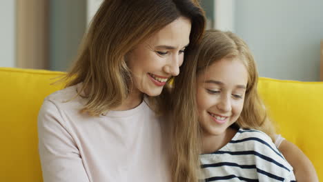 Vista-De-Cerca-De-La-Madre-Y-La-Hija-Rubias-Sonriendo-Y-Viendo-Algo-En-La-Tableta-Mientras-Se-Sientan-En-El-Sofá-En-La-Sala-De-Estar-1