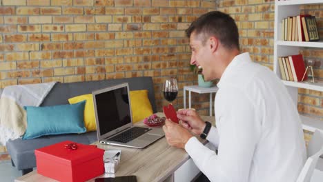 Caucasian-man-making-valentines-day-video-call-using-laptop-holding-red-heart-and-smiling