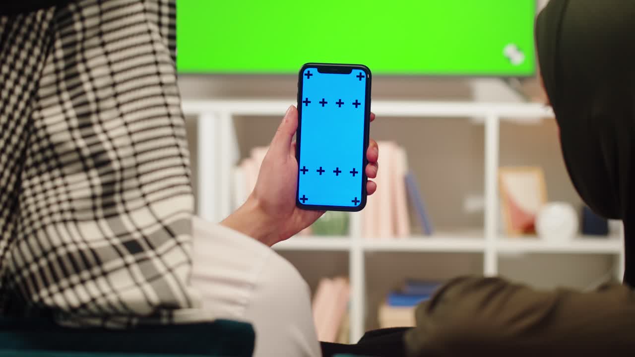 Person Holding Smartphone with Blue Screen in Front of Green Screen TV