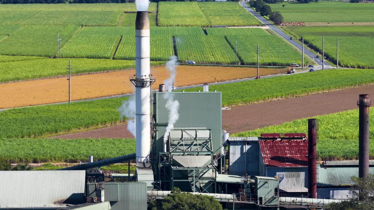 Aerial footage of a sugarcane factory with surrounding fields. Visible smoke emissions and rural landscape under clear daylight