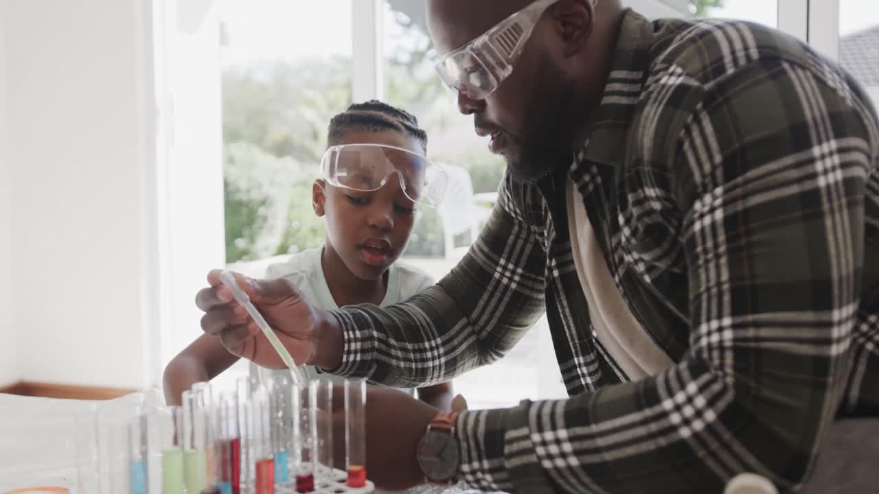 African american father and son sitting at table holding test tubes with liquid, in slow motion